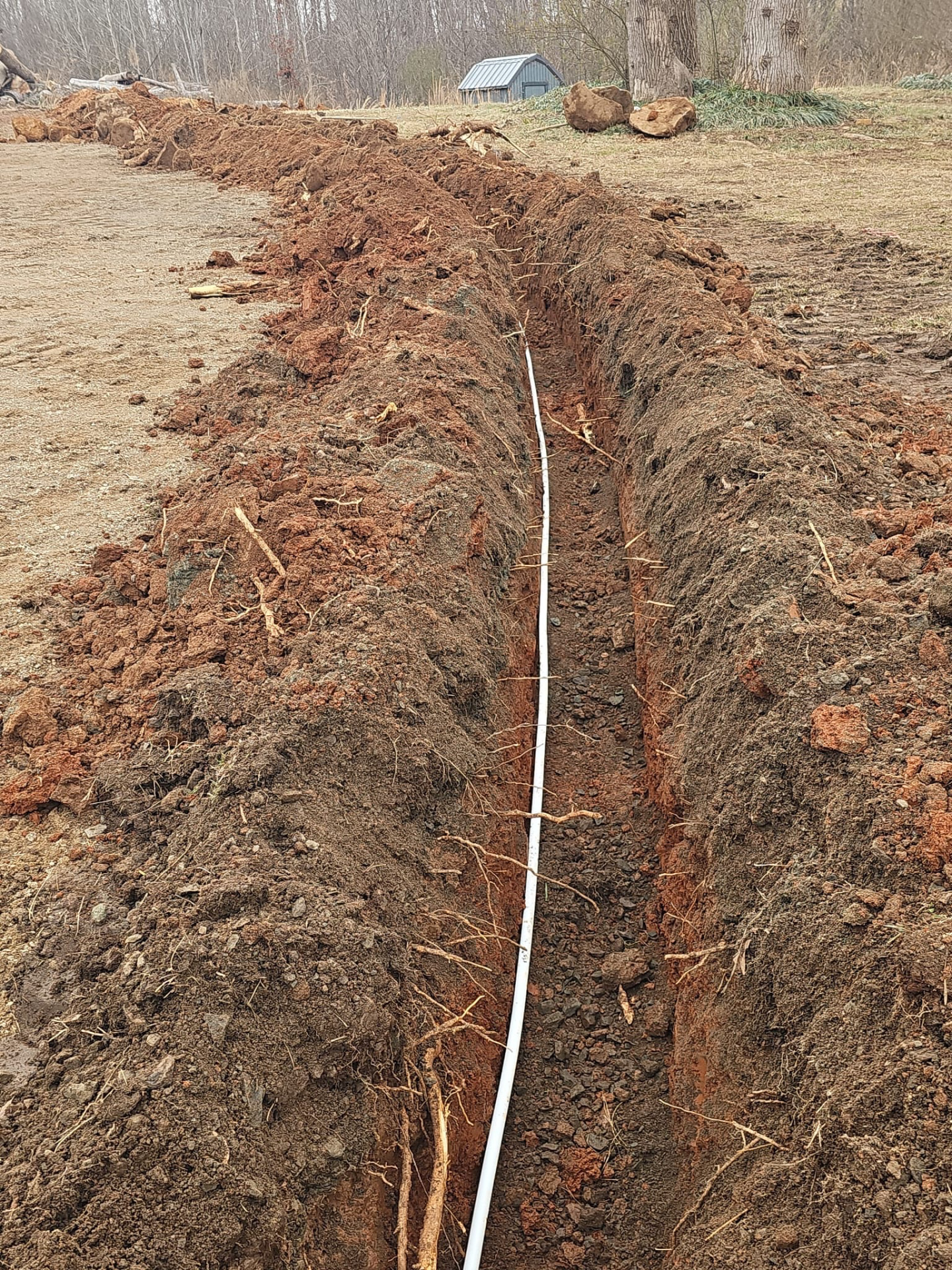 Drainage trench dug through red clay with pipe laid in